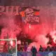 Crowd of football fans in a stadium, red smoke from flares, with a banner featuring a skull and the word 'Ultras' above the goal.