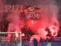Crowd of football fans in a stadium, red smoke from flares, with a banner featuring a skull and the word 'Ultras' above the goal.