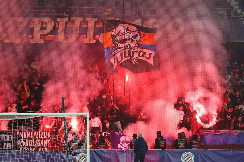 Crowd of football fans in a stadium, red smoke from flares, with a banner featuring a skull and the word 'Ultras' above the goal.