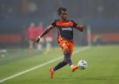 Tchato | AllezPaillade.com Soccer player in orange and navy kit kicking a ball on a green field during a match.