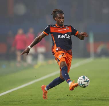 Soccer player in orange and navy kit kicking a ball on a green field during a match.