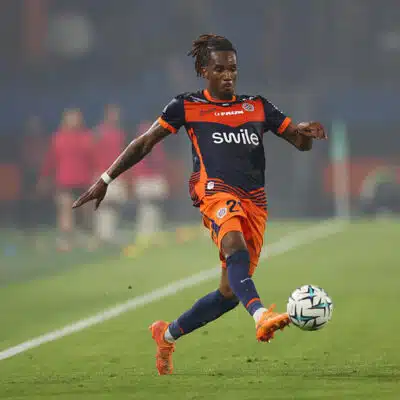 Soccer player in orange and navy kit kicking a ball on a green field during a match.