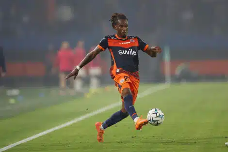 Soccer player in orange and navy kit kicking a ball on a green field during a match.