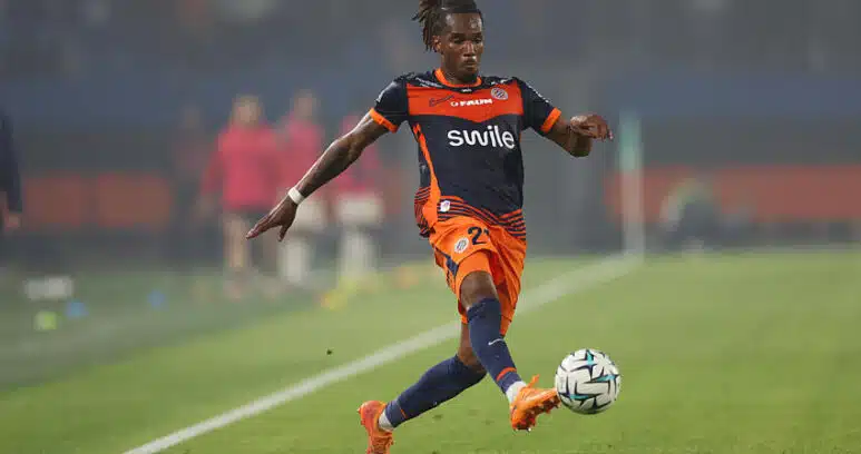 Soccer player in orange and navy kit kicking a ball on a green field during a match.