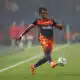 Soccer player in orange and navy kit kicking a ball on a green field during a match.