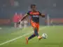 Soccer player in orange and navy kit kicking a ball on a green field during a match.