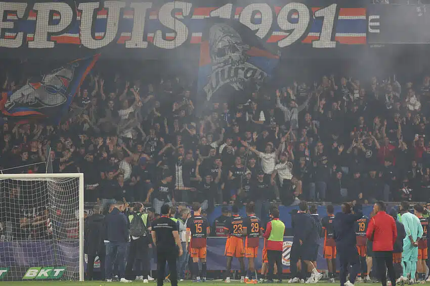 Crowd of football fans behind a goal, with orange-clad players lined up on the sideline.