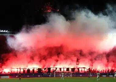 Night stadium scene with fans lighting red flares, creating thick red smoke over the stands.