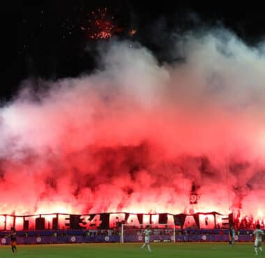 Night stadium scene with fans lighting red flares, creating thick red smoke over the stands.