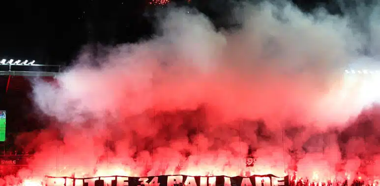 Butte Paillade Ultras fumigène | AllezPaillade.com Night stadium scene with fans lighting red flares, creating thick red smoke over the stands.