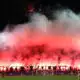 Night stadium scene with fans lighting red flares, creating thick red smoke over the stands.