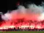 Night stadium scene with fans lighting red flares, creating thick red smoke over the stands.
