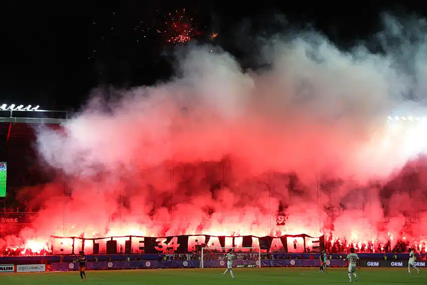 Night stadium scene with fans lighting red flares, creating thick red smoke over the stands.