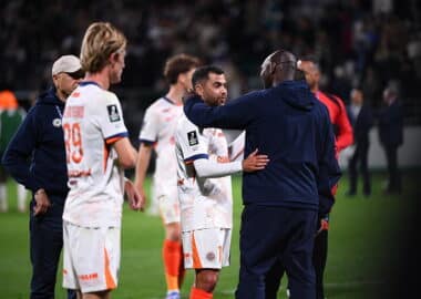 Soccer players on the field share a hug and handshake after a match, with a central player in a white jersey being consoled by a teammate in a dark jacket.