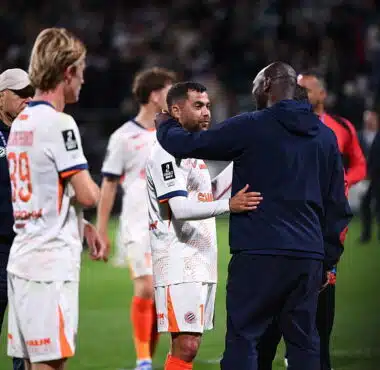 Soccer players on the field share a hug and handshake after a match, with a central player in a white jersey being consoled by a teammate in a dark jacket.