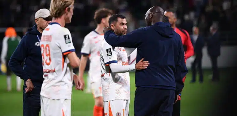 Soccer players on the field share a hug and handshake after a match, with a central player in a white jersey being consoled by a teammate in a dark jacket.