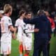 Soccer players on the field share a hug and handshake after a match, with a central player in a white jersey being consoled by a teammate in a dark jacket.