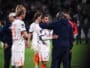 Soccer players on the field share a hug and handshake after a match, with a central player in a white jersey being consoled by a teammate in a dark jacket.