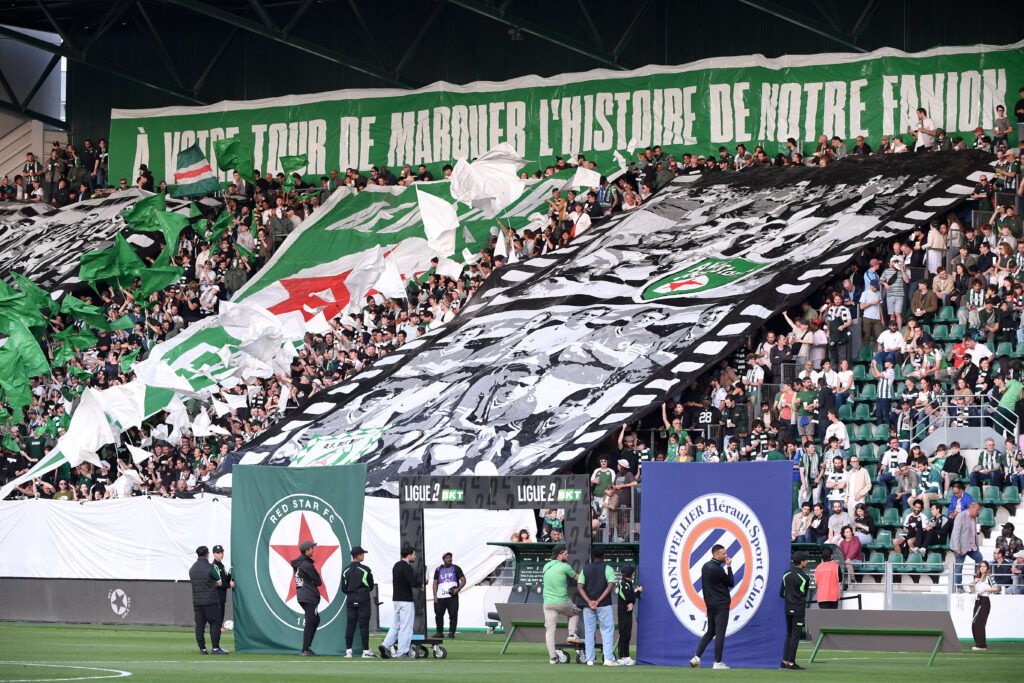 Crowd of soccer fans in a stadium waving green, white, and black banners and flags; a large green banner with white text fills the stands, with security and officials on the sidelines.