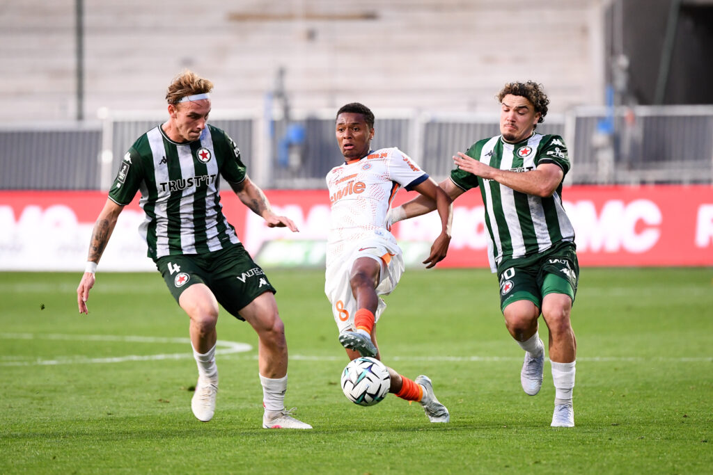 Three soccer players vie for the ball on a grass field, two in green-and-black stripes and one in white and orange kit.