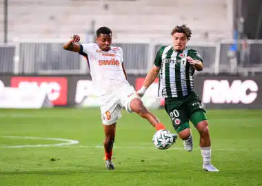 Two soccer players fight for the ball mid‑field: one in white with orange accents (number 8) and the other in green and black stripes, both challenging for possession on the grass field