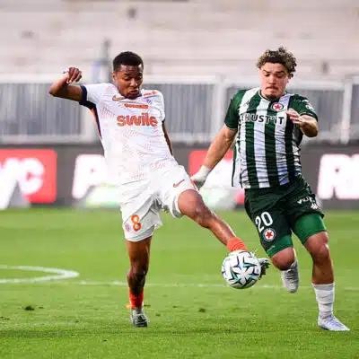 Two soccer players fight for the ball mid‑field: one in white with orange accents (number 8) and the other in green and black stripes, both challenging for possession on the grass field