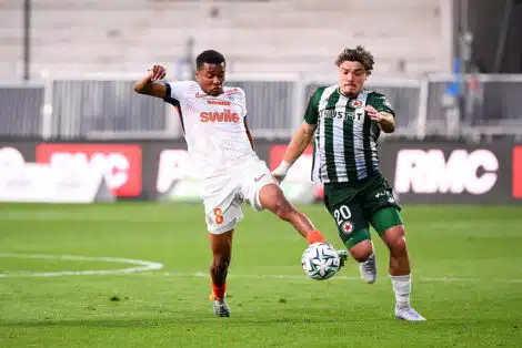 Two soccer players fight for the ball mid‑field: one in white with orange accents (number 8) and the other in green and black stripes, both challenging for possession on the grass field