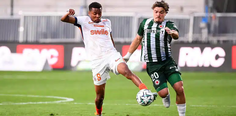 Two soccer players fight for the ball mid‑field: one in white with orange accents (number 8) and the other in green and black stripes, both challenging for possession on the grass field