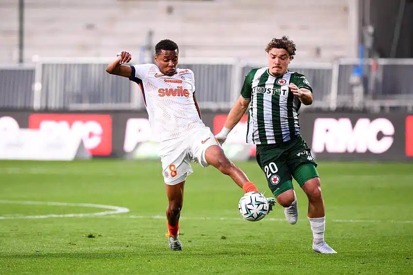 Two soccer players fight for the ball mid‑field: one in white with orange accents (number 8) and the other in green and black stripes, both challenging for possession on the grass field