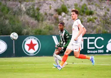 Two soccer players sprint after the ball on a green field, one in green-and-black stripes and the other in white with orange socks.",