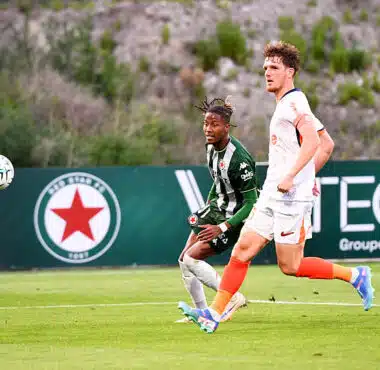 Two soccer players sprint after the ball on a green field, one in green-and-black stripes and the other in white with orange socks.",