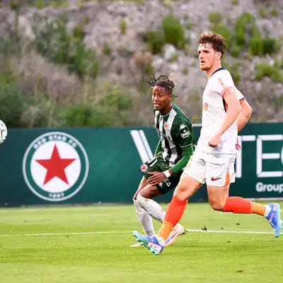 Two soccer players sprint after the ball on a green field, one in green-and-black stripes and the other in white with orange socks.",