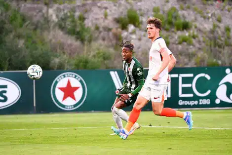 Two soccer players sprint after the ball on a green field, one in green-and-black stripes and the other in white with orange socks.",