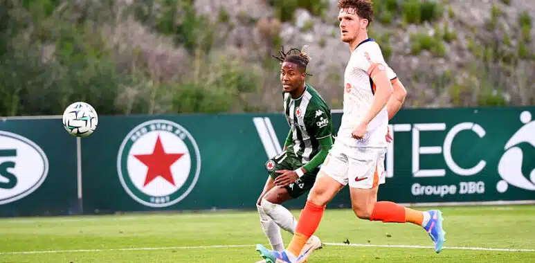 Two soccer players sprint after the ball on a green field, one in green-and-black stripes and the other in white with orange socks.",