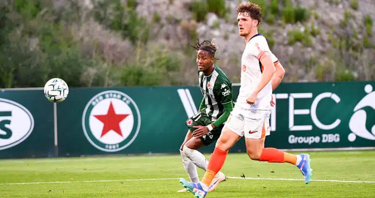 Two soccer players sprint after the ball on a green field, one in green-and-black stripes and the other in white with orange socks.",