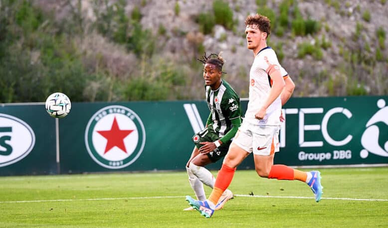 Two soccer players sprint after the ball on a green field, one in green-and-black stripes and the other in white with orange socks.",
