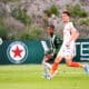 Two soccer players sprint after the ball on a green field, one in green-and-black stripes and the other in white with orange socks.",