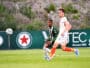 Two soccer players sprint after the ball on a green field, one in green-and-black stripes and the other in white with orange socks.",
