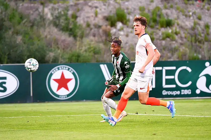 Two soccer players sprint after the ball on a green field, one in green-and-black stripes and the other in white with orange socks.",