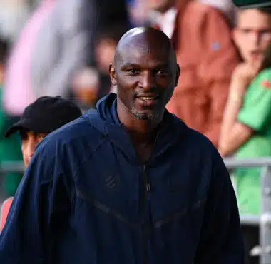 Man in a navy jacket smiles as he stands at an outdoor event, with blurred crowd members in the background.