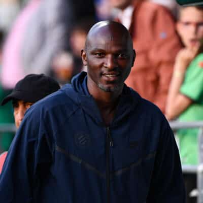 Man in a navy jacket smiles as he stands at an outdoor event, with blurred crowd members in the background.