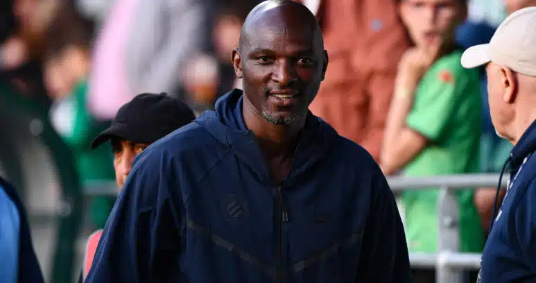 Man in a navy jacket smiles as he stands at an outdoor event, with blurred crowd members in the background.