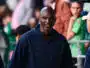 Man in a navy jacket smiles as he stands at an outdoor event, with blurred crowd members in the background.
