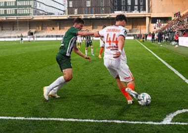 Two soccer players contest the ball near the sideline on a green pitch as a white jersey #44 (Chennai) clashes with a green opponent, with stands and crowd in the background.
