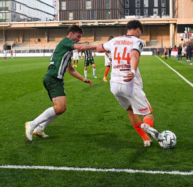 Two soccer players contest the ball near the sideline on a green pitch as a white jersey #44 (Chennai) clashes with a green opponent, with stands and crowd in the background.