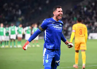 Maubleu | AllezPaillade.com Soccer goalkeeper in a blue kit celebrates with arms outstretched on the field, green-clad players blurred in the background.