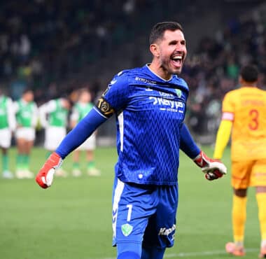 Soccer goalkeeper in a blue kit celebrates with arms outstretched on the field, green-clad players blurred in the background.