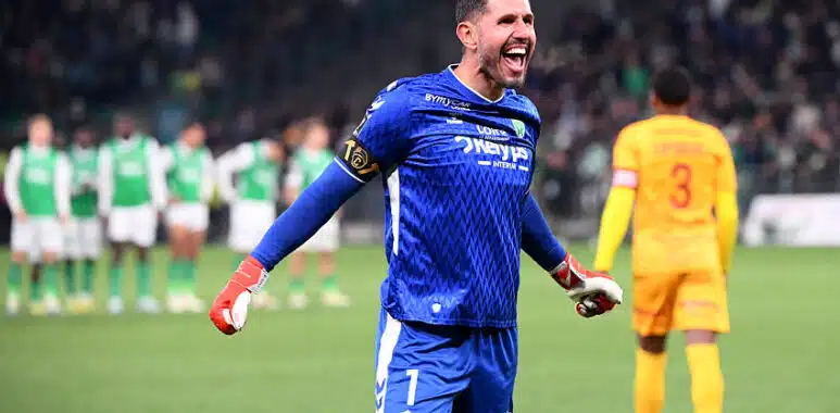 Soccer goalkeeper in a blue kit celebrates with arms outstretched on the field, green-clad players blurred in the background.