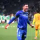 Soccer goalkeeper in a blue kit celebrates with arms outstretched on the field, green-clad players blurred in the background.