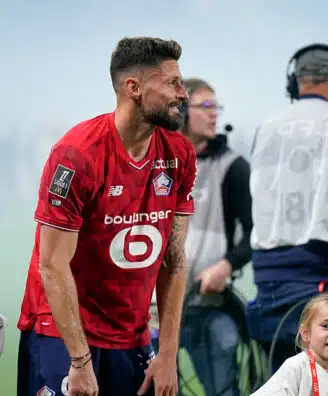Bearded soccer player in a red jersey bends slightly forward on a field, surrounded by children and a filming crew in the background.